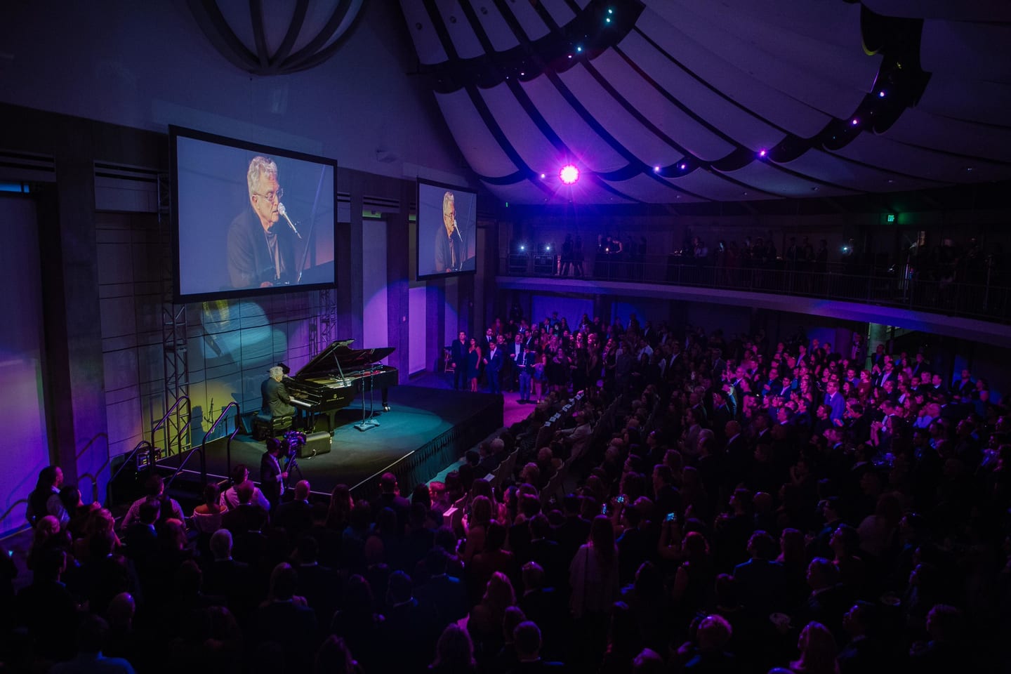 Randy Newman playing a grand piano on a darkened stage under blue light before a full house