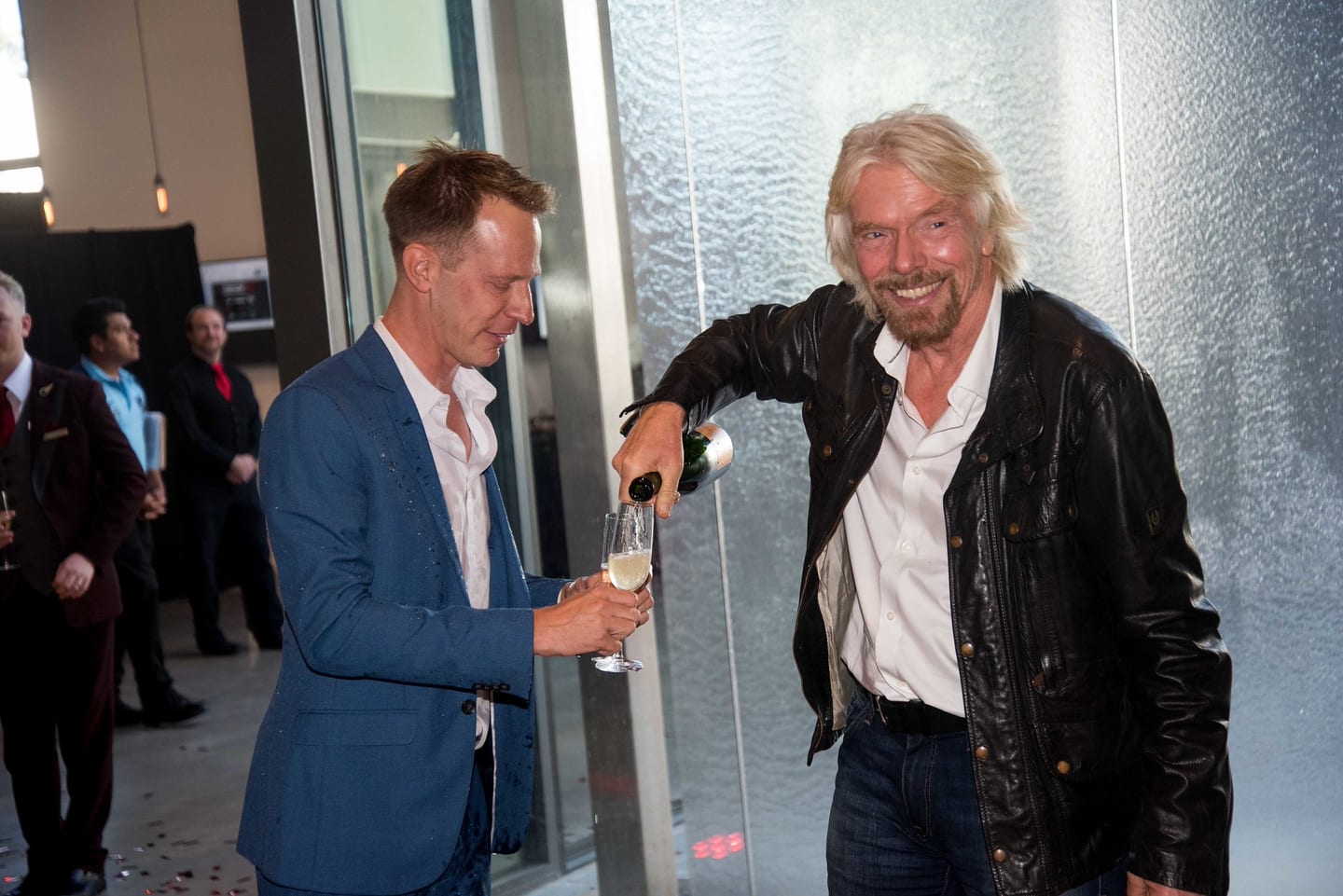 Richard Branson laughing as he pours a glass of champagne for a man in a blue suit at a Virgin event
