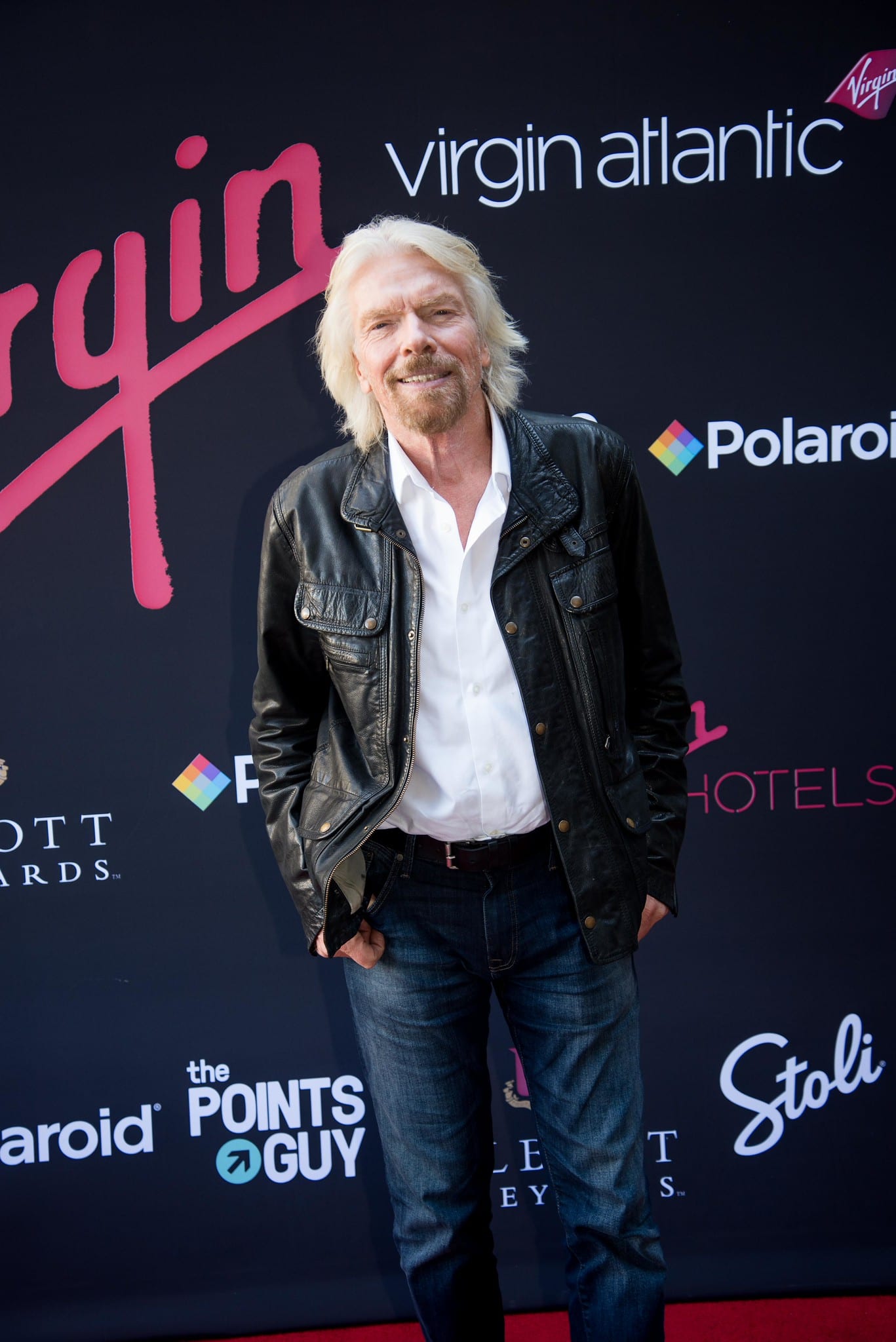 Richard Branson in a leather jacket on the red carpet in front of a Virgin Atlantic step-and-repeat