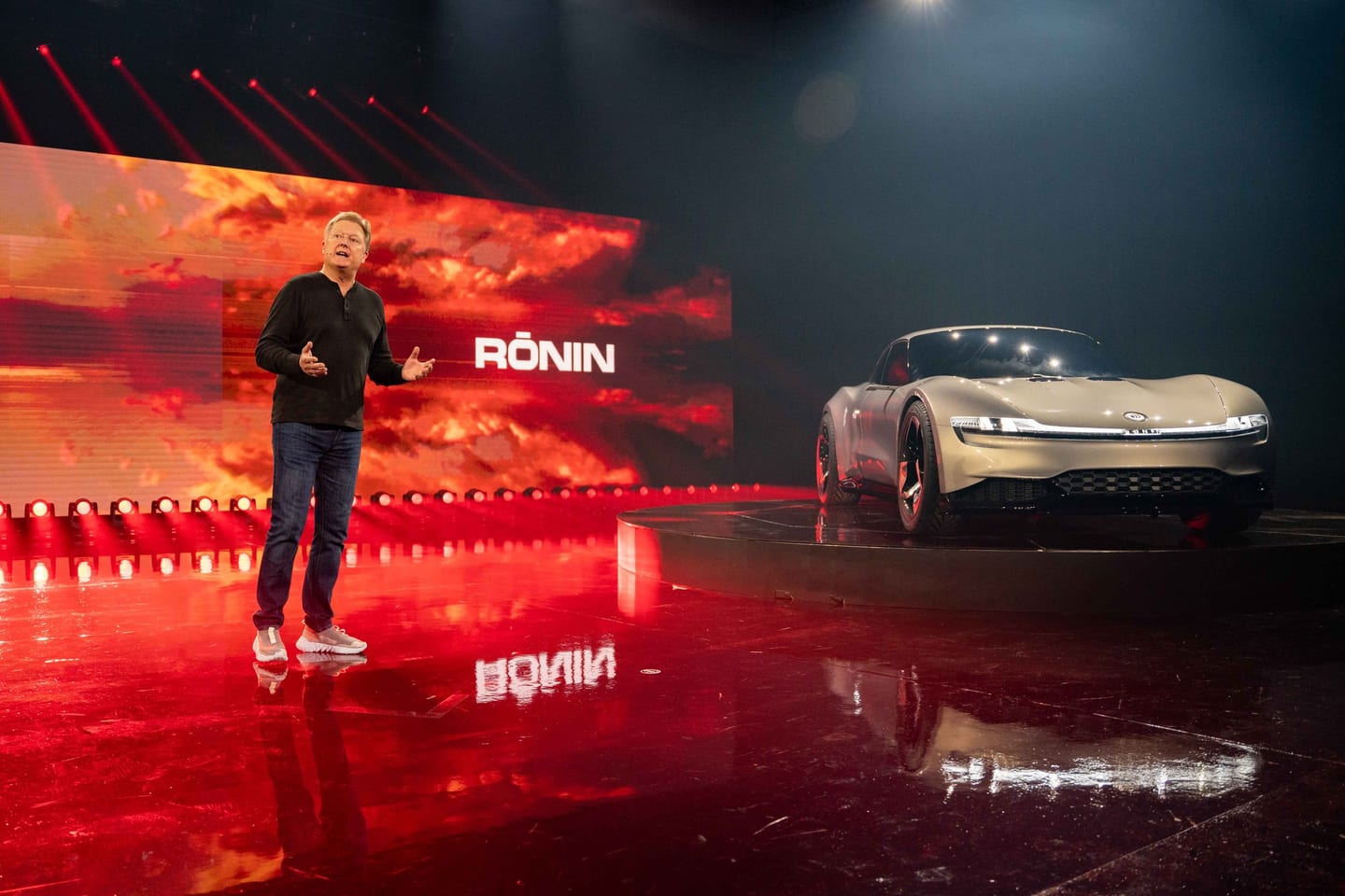 A presenter on stage gesturing beside the silver Fisker Ronin on a rotating platform against a red screen reading Ronin