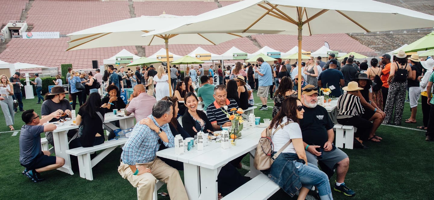 Festival-goers seated at picnic tables under umbrellas among vendor tents on the Rose Bowl field, the Pasadena Tournament of Roses banner behind