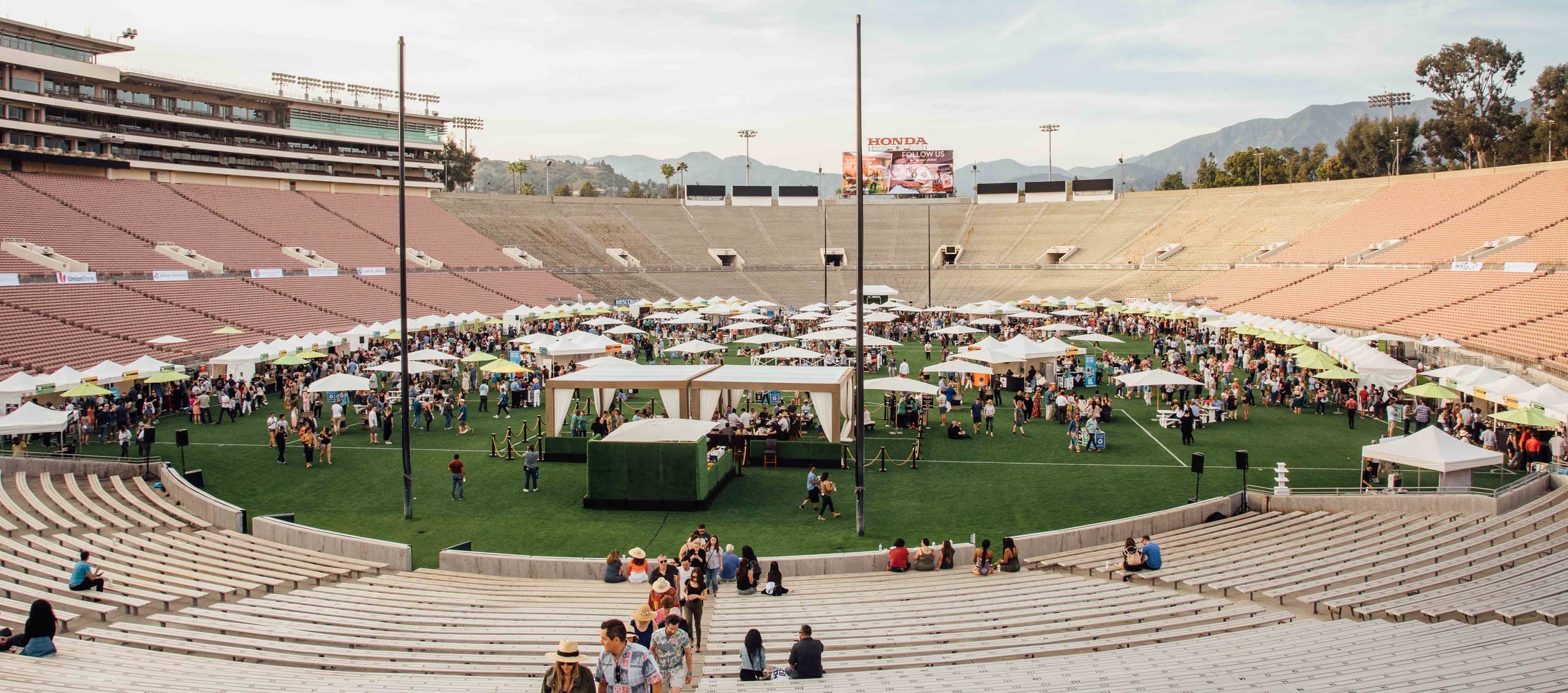 A food and drink festival filling the field of the Rose Bowl stadium in Pasadena, rows of vendor tents across the turf with the grandstands and San Gabriel mountains behind at golden hour