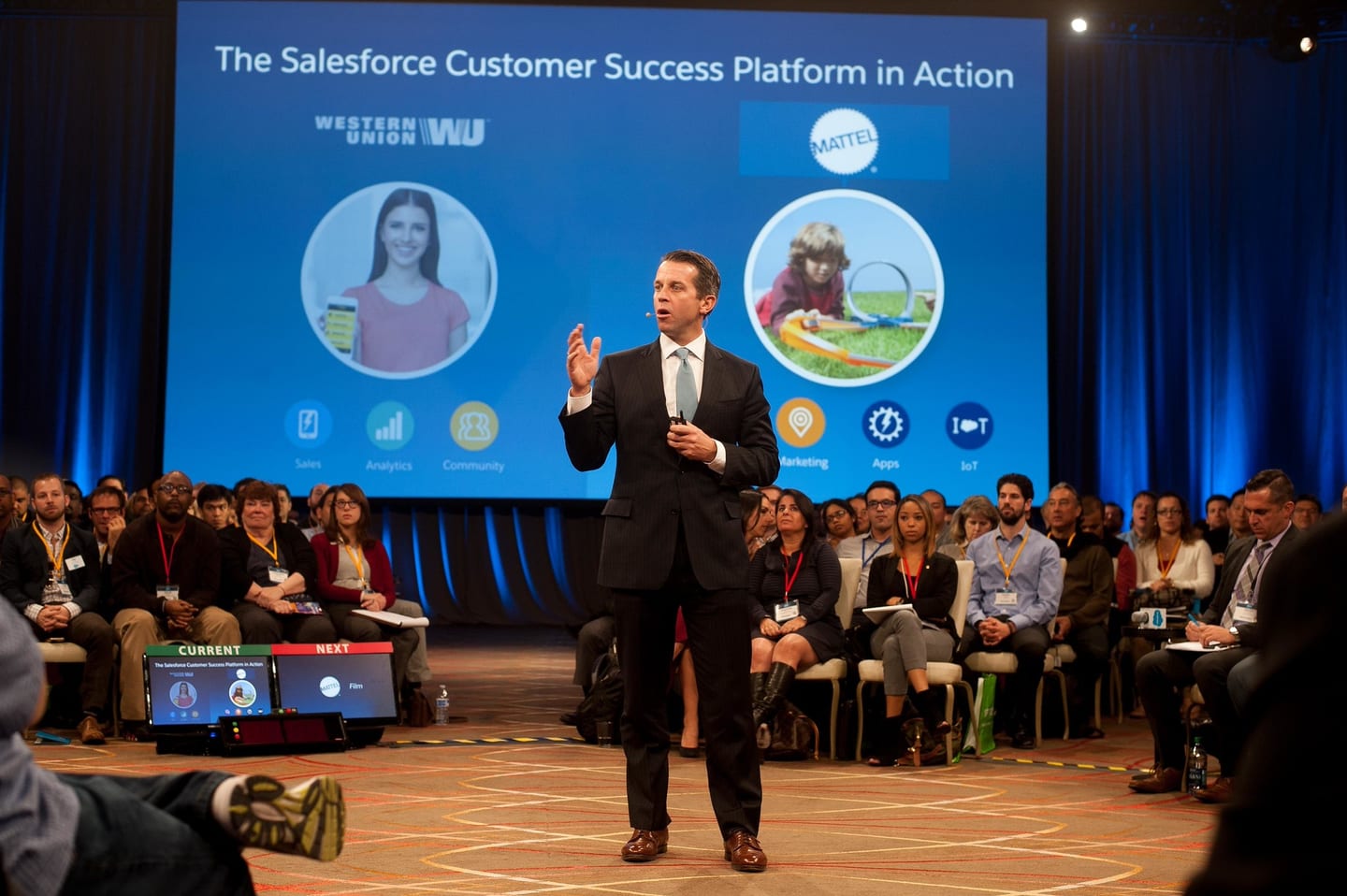 A presenter on stage before a large screen at a Salesforce keynote with a seated audience