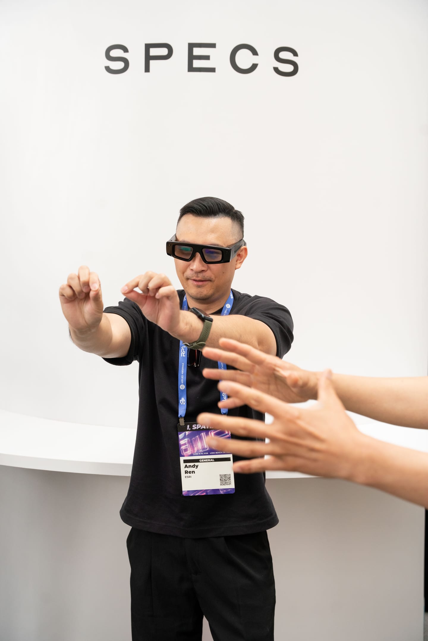An attendee wearing Snap Specs reaching both hands forward toward another person’s hands during a hand-tracking demo under a SPECS sign