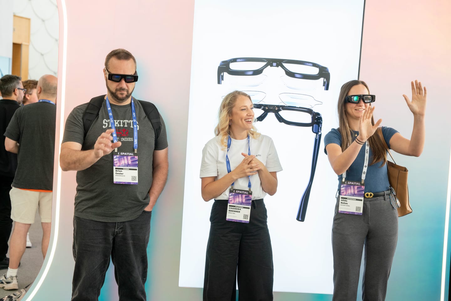 Three attendees wearing Snap Specs gesturing beside a large backlit product image of the glasses