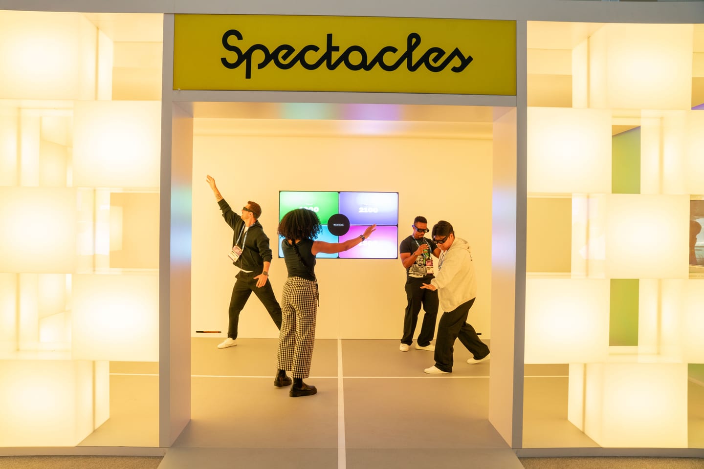 Four attendees wearing Snap Spectacles gesturing in mid-air inside a demo room lined with glowing white cubes, beneath a Spectacles sign