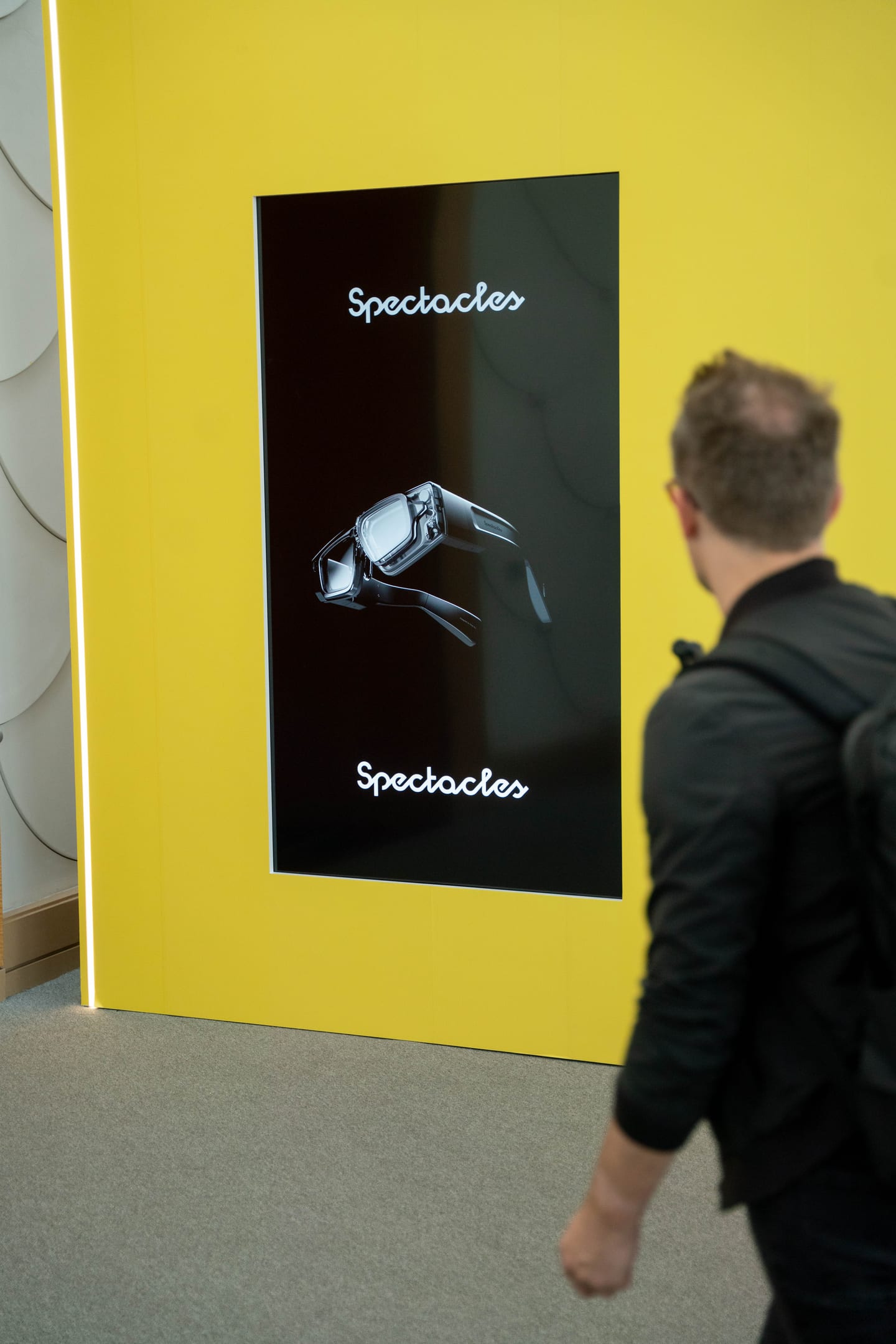 A man carrying a backpack walking past a vertical screen showing Snap Spectacles against a bright yellow wall