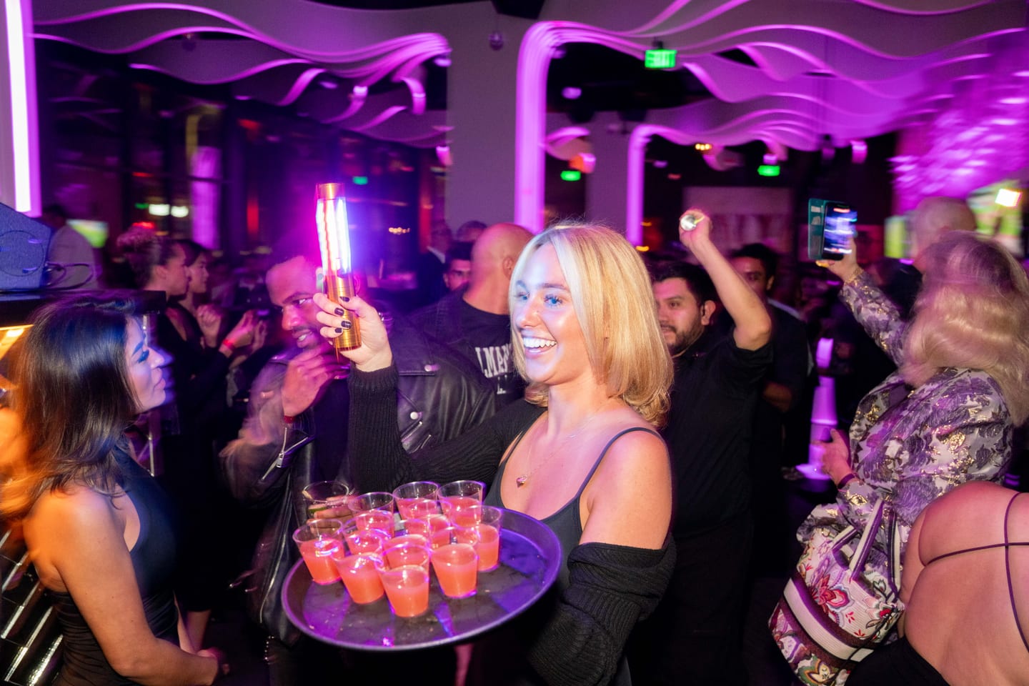 A server carrying a tray of pink shots through a purple-lit party crowd at STK Westwood