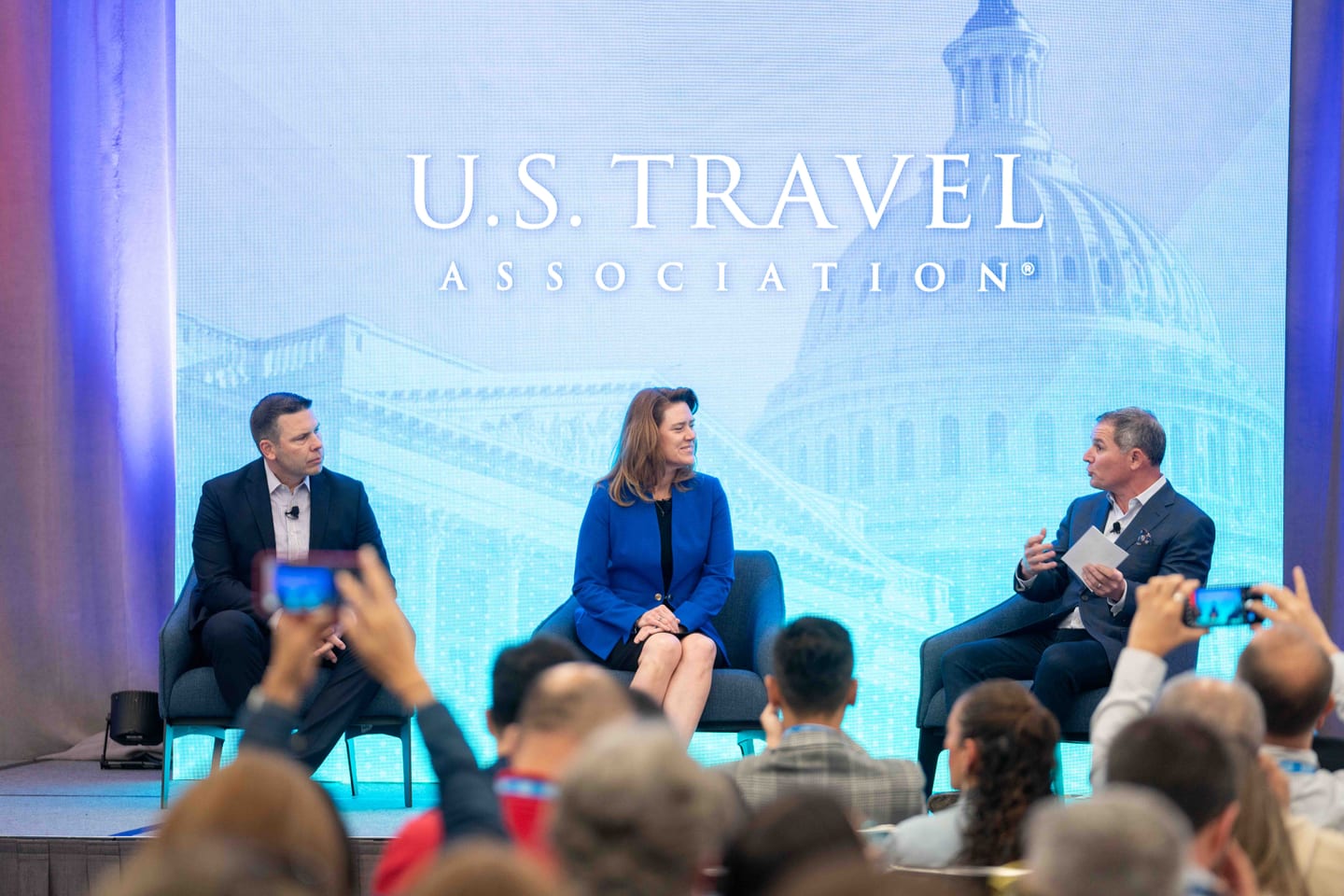 Three panelists seated on stage at the U.S. Travel Association press conference before a screen showing the U.S. Capitol