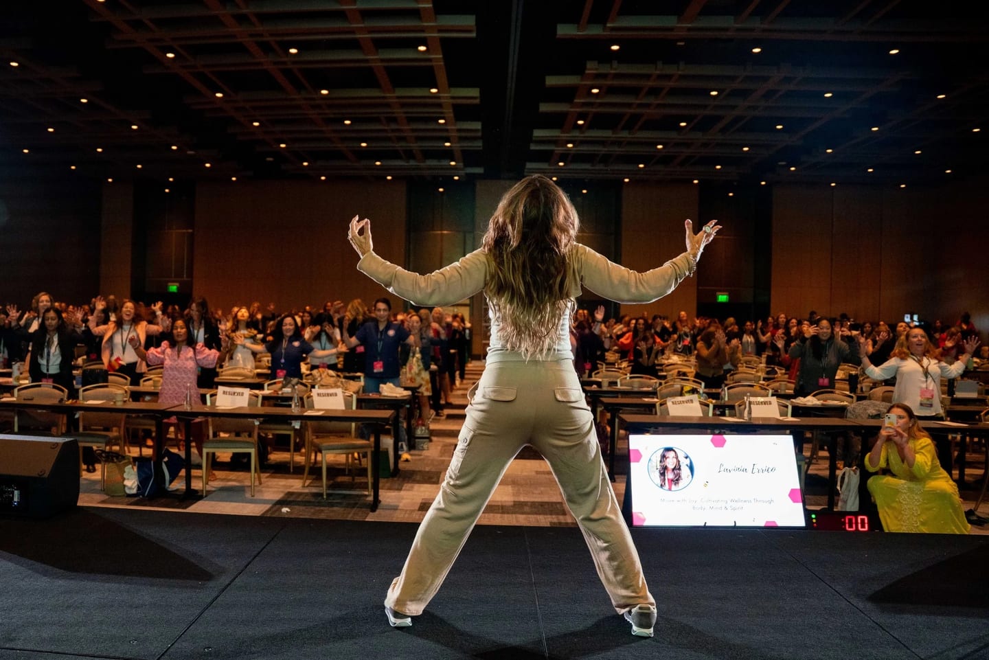 A speaker with arms raised leading a packed ballroom of attendees on their feet at a Los Angeles wellness summit