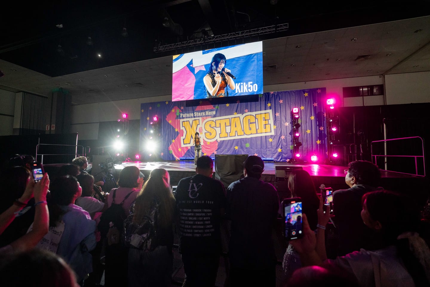 A crowd holding up phones to film a performer on the X Stage at KCON LA 2025, with the artist shown on the screen overhead