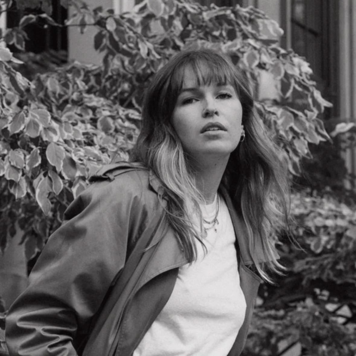 A woman with wavy hair and bangs, wearing a jacket over a T-shirt, standing outdoors near leafy bushes
